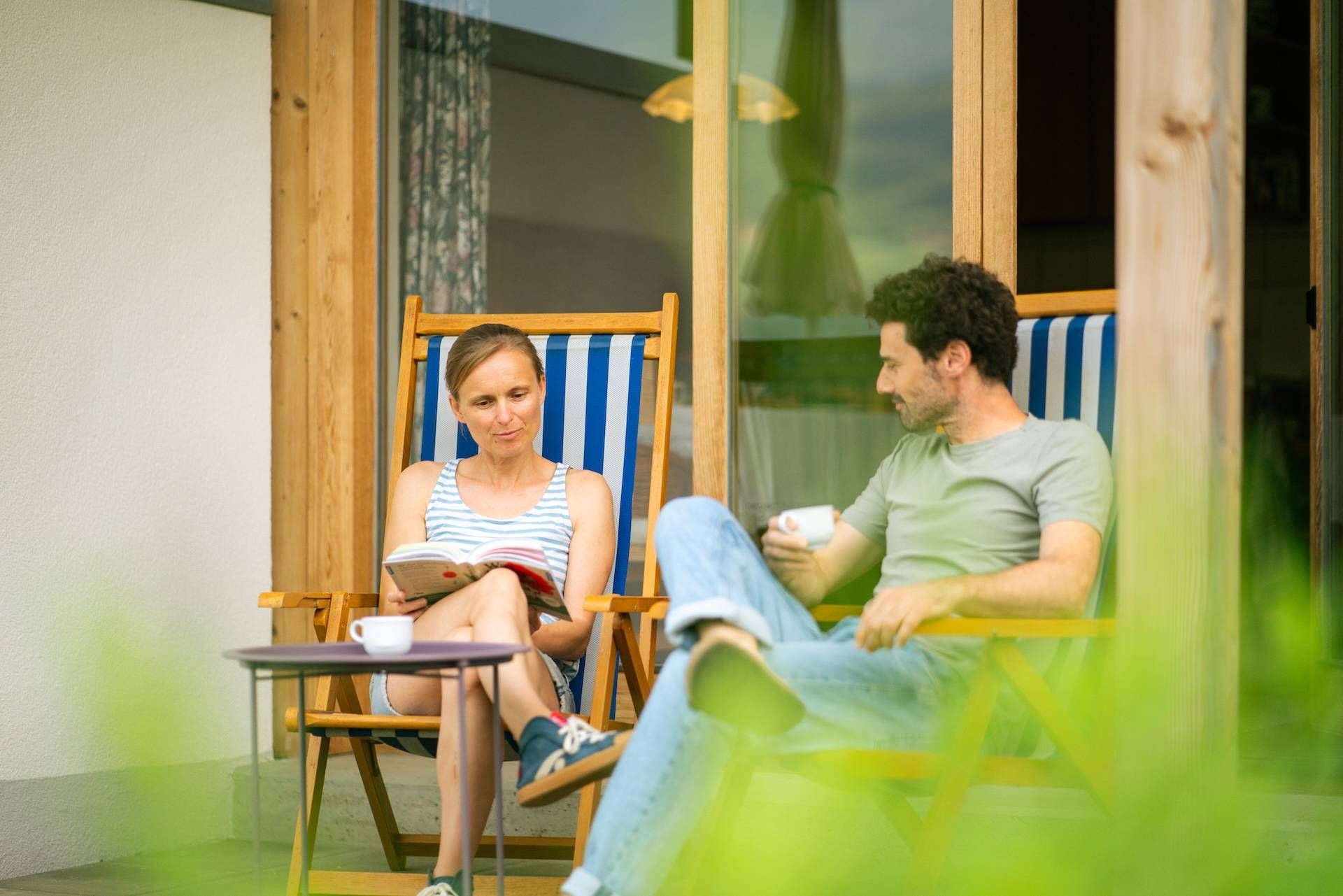 Zwei Personen sitzen auf Sonnenstühlen und die Frau liest ein Buch und der Mann trinkt einen Kaffee