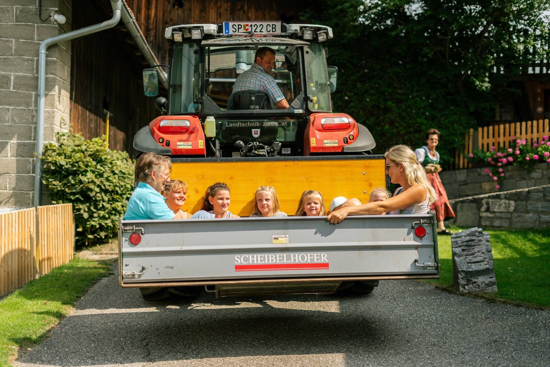 Kinder und Erwachsene sitzen im Hänger eines Traktors und fahren über den Hof
