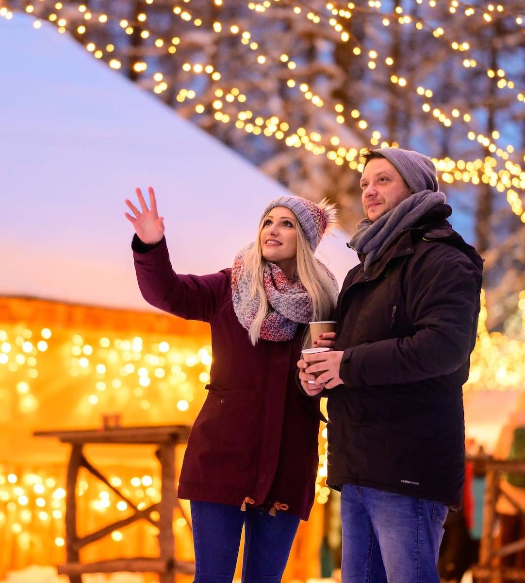 Junges Paar beim Christkindlmarkt mit Lichterketten und schneebedeckten Dächern im Hintergrund